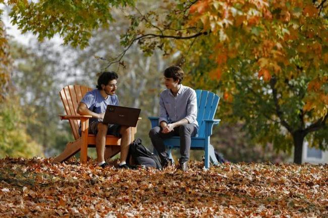 two students sitting in colorful Adirondack chairs with fall colored leaves surrounding them 